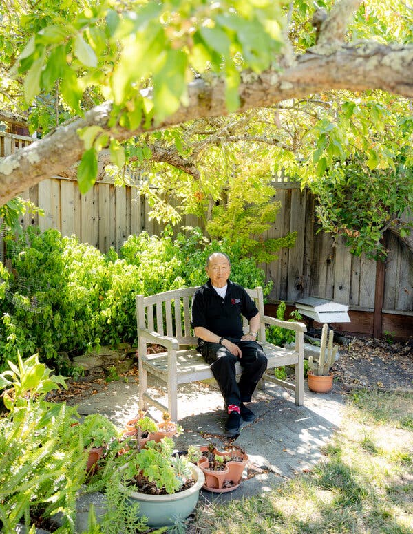 Fukuyama sits on a wooden bench in a garden, surrounded by green foliage and potted plants. He wears a black shirt and dark pants.