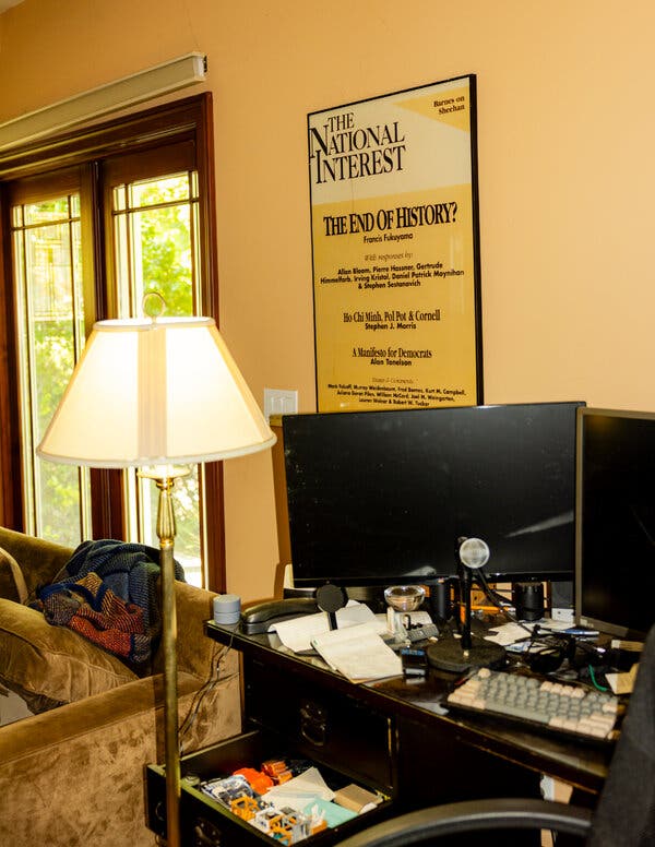 A room with a floor lamp, a brown sofa and a cluttered dark desk with monitors. A poster on a yellow wall reads "THE NATIONAL INTEREST" and "THE END OF HISTORY?"